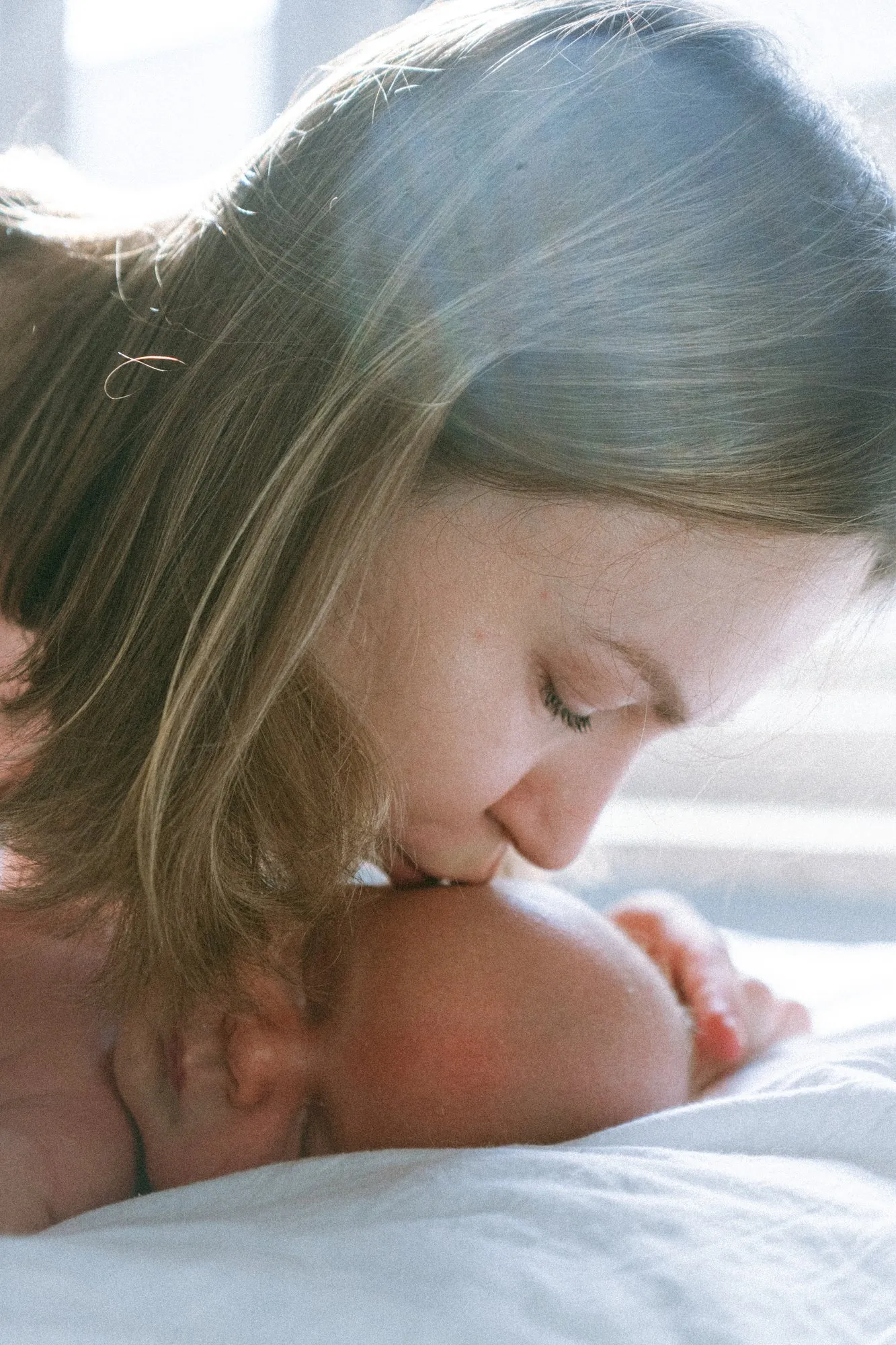 Mama całuje śpiącego noworodka w główkę, miękkie światło z okna.