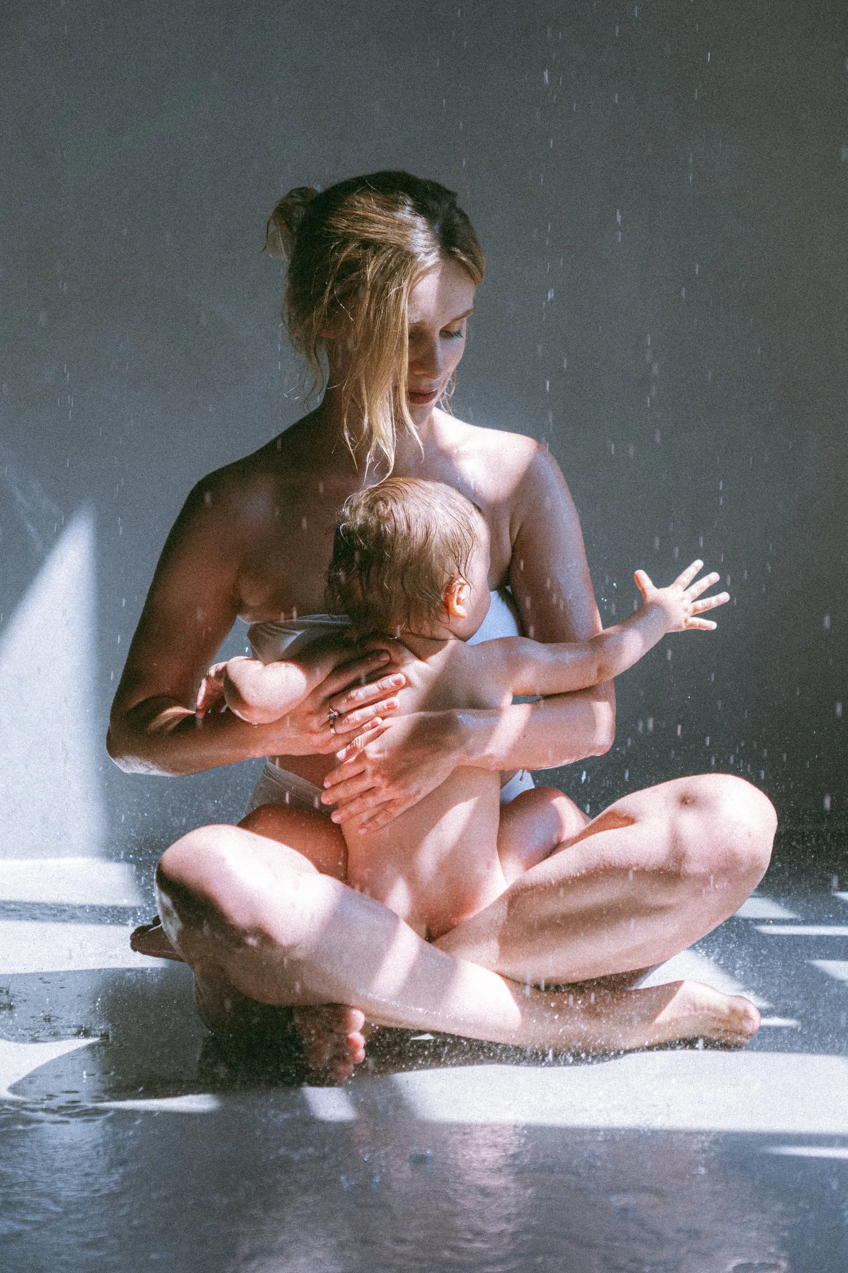 Mama siedzi po turecku pod deszczownicą z niemowlęciem w ramionach, wokół krople wody.