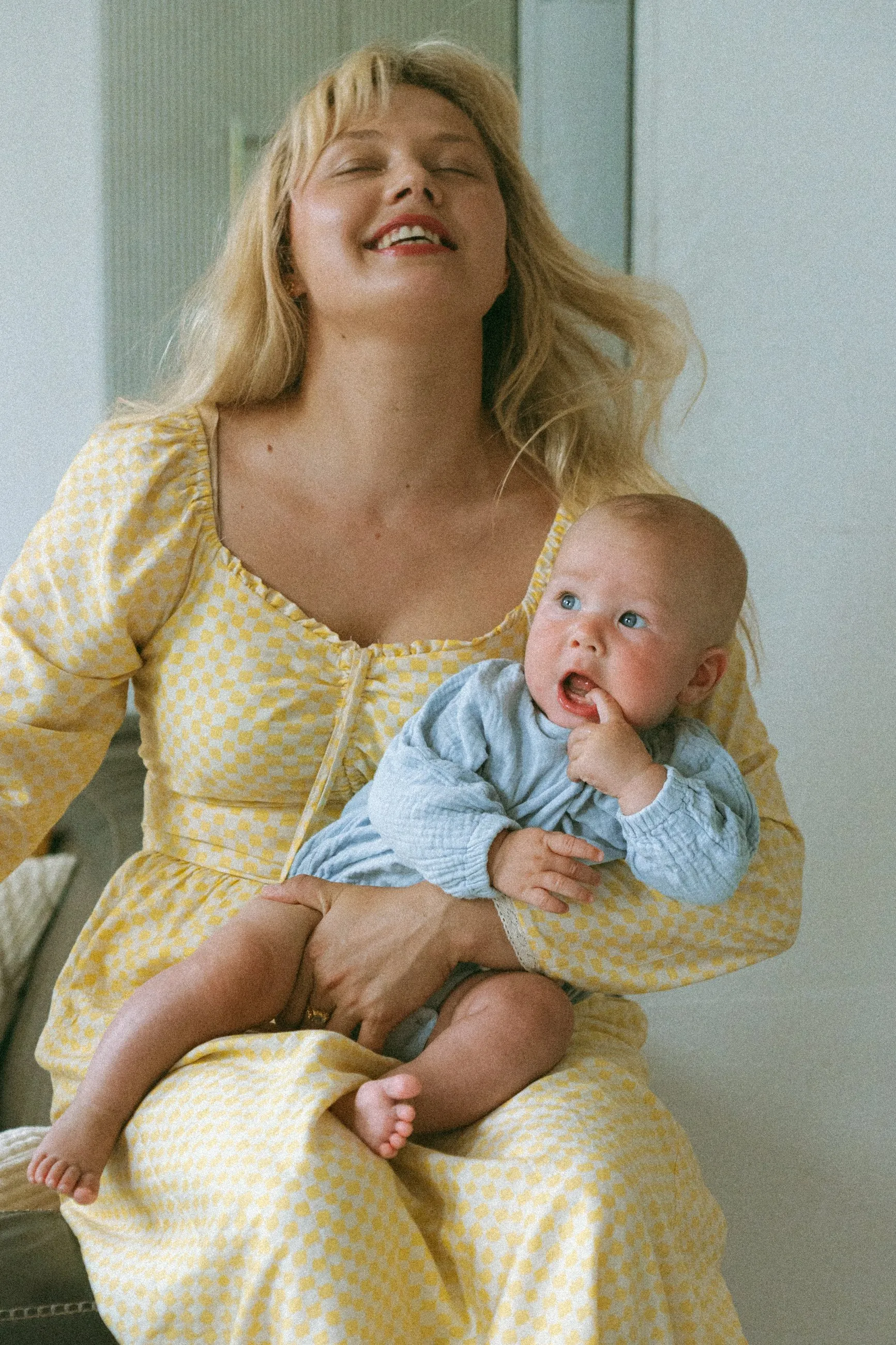 Mama w żółtej sukience śmieje się z zamkniętymi oczami, niemowlę na rękach patrzy w bok.